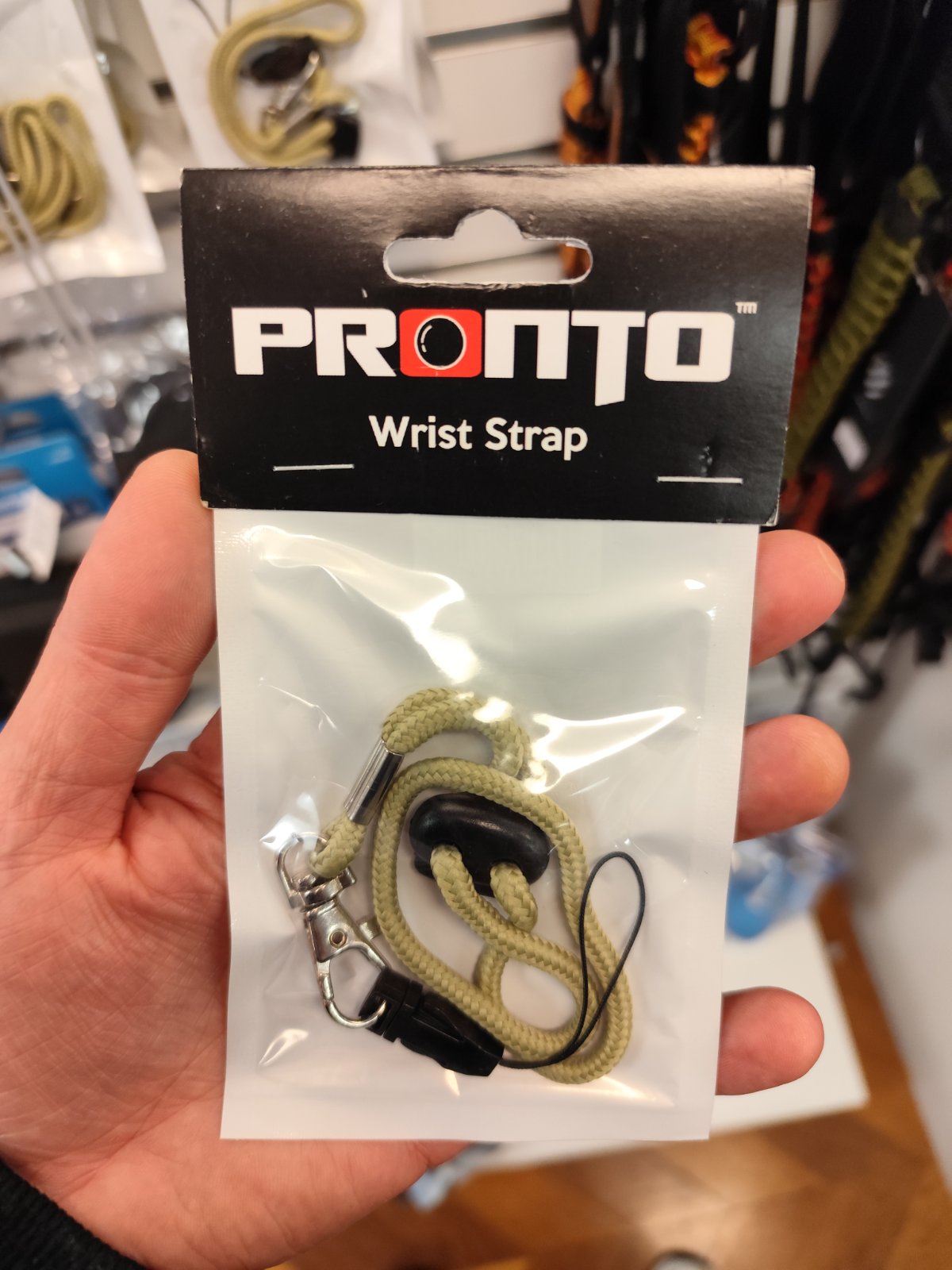 Hand holding a packaged Pronto wrist strap with beige cord and clip, shop shelves behind