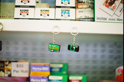 Two keychains resembling film packaging on a shelf with photography supplies in the background.